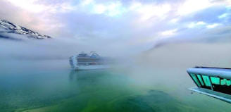 A large cruise ship is partially obscured by dense fog on a body of water. The surrounding landscape includes snow-topped mountains and a cloudy sky. In the foreground, part of a ship’s deck with reflective windows is visible.