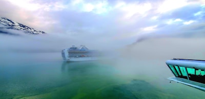A large cruise ship is partially obscured by dense fog on a body of water. The surrounding landscape includes snow-topped mountains and a cloudy sky. In the foreground, part of a ship’s deck with reflective windows is visible.