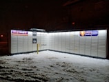 A parcel locker unit illuminated at night, surrounded by snow. The lockers display various advertising signs and logos, including 'InPost' and 'allegro SMART', and are accessible 24/7 as indicated by the signage.