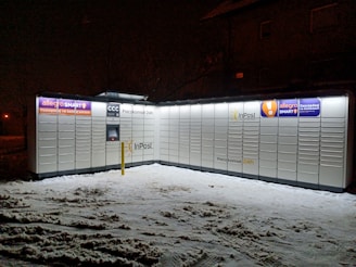 A parcel locker unit illuminated at night, surrounded by snow. The lockers display various advertising signs and logos, including 'InPost' and 'allegro SMART', and are accessible 24/7 as indicated by the signage.