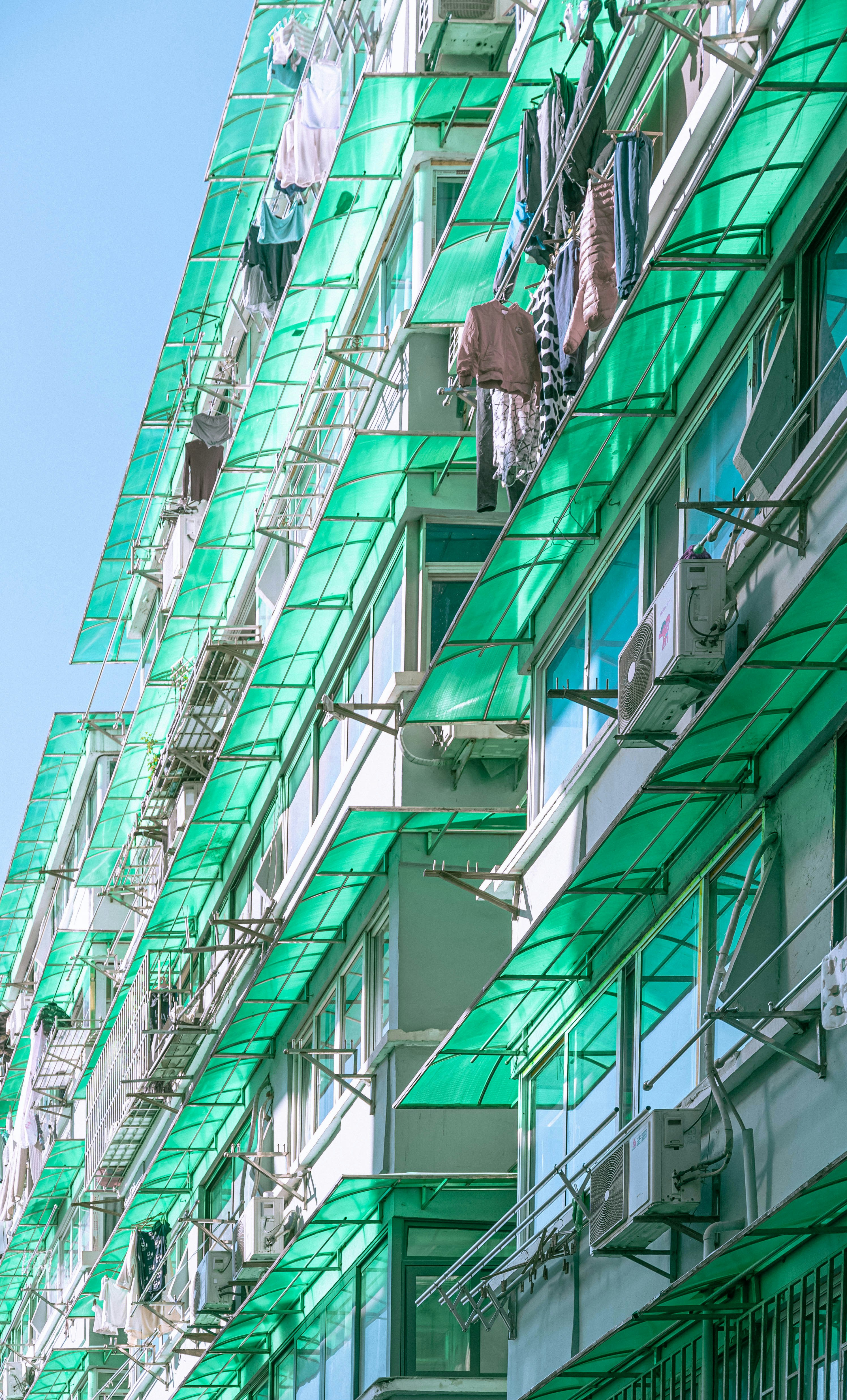 A residential building with multiple floors featuring green-tinted glass balconies and awnings. Clothes and laundry are hung out to dry on lines extending from the balconies. Air conditioning units are also visible on the walls.