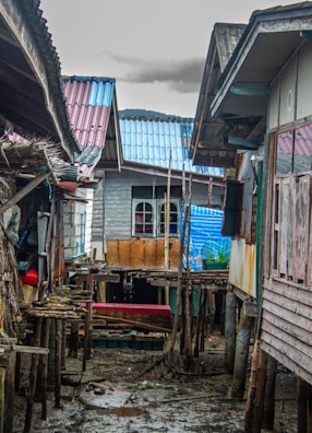 Several wooden stilt houses with corrugated metal roofs, weathered by time, are built closely together. The narrow pathway between the houses is muddy and littered with debris. Materials such as planks, tarpaulins, and makeshift structures are visible, indicating a humble and rustic living environment. The sky above is overcast, contributing to a somber mood.