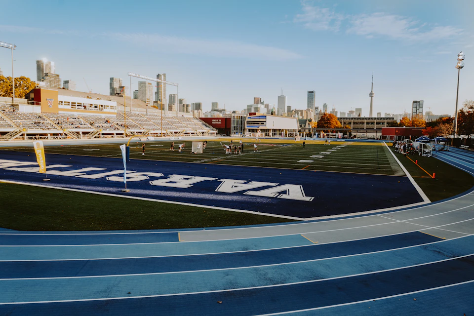The image shows a large, empty stadium with a blue and white color scheme. The field is marked by white lines that define its boundaries, and there are orange cones scattered around the perimeter. In the background, the city skyline can be seen, featuring tall buildings of varying heights against a clear sky.