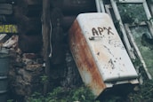 A rusty, old refrigerator with graffiti is leaning against the exterior of a rundown wooden structure. Surrounding the refrigerator are broken windows, overgrown plants, and weathered stone and wood surfaces. There is a sense of neglect and decay.