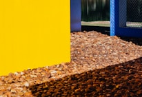 A bright yellow wall intersects with a blue chain-link fence. The ground is covered with brown, tan, and gray pebbles, creating a textured foreground. In the background, there's a glimpse of a green lawn and a body of water reflecting light.