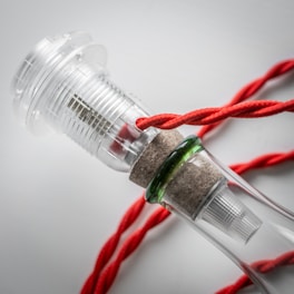 A close-up view of a modern, transparent bottle with a cork. The bottle has a unique design featuring a section of clear plastic or glass, encapsulating a red rope that is artistically wrapped around it. The photograph captures the intricate textures of the materials, emphasizing the glossy surface of the bottle and the fibrous texture of the rope.