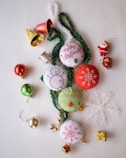 A collection of Christmas-themed decorations including fabric ornaments with embroidered designs like snowflakes and a snowman, green tinsel, shiny baubles in red and green, small gold-wrapped gifts, a decorative Santa figure, small bells, and a translucent snowflake.