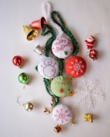 A collection of Christmas-themed decorations including fabric ornaments with embroidered designs like snowflakes and a snowman, green tinsel, shiny baubles in red and green, small gold-wrapped gifts, a decorative Santa figure, small bells, and a translucent snowflake.