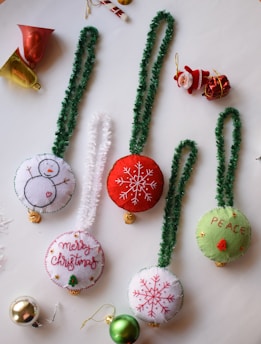A collection of festive Christmas decorations featuring handmade plush ornaments with embroidered designs including snowflakes, a snowman, the word 'PEACE', and 'Merry Christmas'. There are different colored bell ornaments and a small Santa Claus figure nearby. The ornaments have green and white fluffy hangers, and additional decorations like a red and gold metallic bell, a candy cane, and Christmas balls are scattered around.