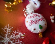 A handmade Christmas ornament lies on a festive red surface. The ornament is circular, with white fabric stitched with red thread spelling 'Merry Christmas'. Small decorative elements include a green Christmas tree and tiny stars. In the background, golden bells and a translucent snowflake decoration are visible.
