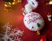 A handmade Christmas ornament lies on a festive red surface. The ornament is circular, with white fabric stitched with red thread spelling 'Merry Christmas'. Small decorative elements include a green Christmas tree and tiny stars. In the background, golden bells and a translucent snowflake decoration are visible.