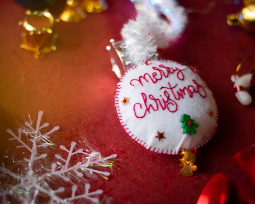 A handmade Christmas ornament lies on a festive red surface. The ornament is circular, with white fabric stitched with red thread spelling 'Merry Christmas'. Small decorative elements include a green Christmas tree and tiny stars. In the background, golden bells and a translucent snowflake decoration are visible.