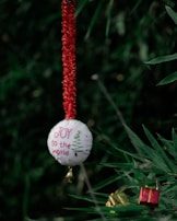A circular white ornament with red stitching hangs from a textured, red string. The ornament features embroidered text 'JOY to the world' alongside a simple green Christmas tree design. A small golden bell is attached to the bottom of the ornament. In the background, there are blurred green leaves and a pair of small, wrapped gifts in red and green.