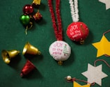 Festive decorations are arranged on a green textured surface. The items include red and white ornaments with 'JOY to the world' embroidered alongside a Christmas tree design, shiny golden and red bells, small wrapped gift boxes, and cut-out stars in silver and gold. Red and white tinsel strings accompany the ornaments, adding a cheerful and celebratory touch.