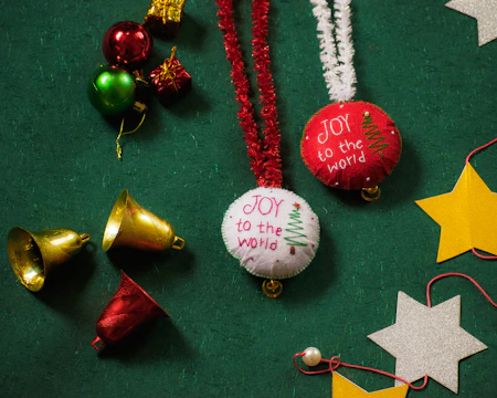 Festive decorations are arranged on a green textured surface. The items include red and white ornaments with 'JOY to the world' embroidered alongside a Christmas tree design, shiny golden and red bells, small wrapped gift boxes, and cut-out stars in silver and gold. Red and white tinsel strings accompany the ornaments, adding a cheerful and celebratory touch.