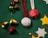 Festive decorations are arranged on a green textured surface. The items include red and white ornaments with 'JOY to the world' embroidered alongside a Christmas tree design, shiny golden and red bells, small wrapped gift boxes, and cut-out stars in silver and gold. Red and white tinsel strings accompany the ornaments, adding a cheerful and celebratory touch.