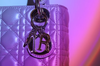 A close-up view of a quilted leather bag with metallic hardware, including a shiny metallic charm with the letters 'D' and 'R'. The bag's surface has a reflective texture that catches light, creating a vibrant, colorful gradient effect of purple and pink tones.