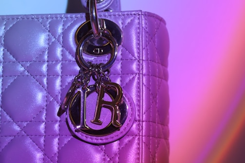 A close-up view of a quilted leather bag with metallic hardware, including a shiny metallic charm with the letters 'D' and 'R'. The bag's surface has a reflective texture that catches light, creating a vibrant, colorful gradient effect of purple and pink tones.