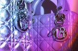 Two luxurious handbags with textured, quilted patterns are adorned with shiny metal charms. The lighting casts shades of purple and pink, creating a vibrant effect on the metallic surfaces.