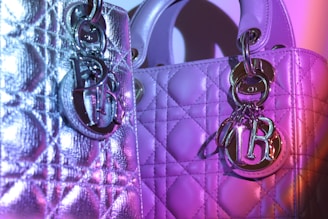 Two luxurious handbags with textured, quilted patterns are adorned with shiny metal charms. The lighting casts shades of purple and pink, creating a vibrant effect on the metallic surfaces.