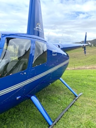 A bright blue helicopter with the words 'Sydney Heli Tours' is parked on a grassy field. The helicopter model is R44 Raven II. The sky is overcast with scattered clouds.