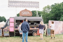 A black food truck adorned with colorful patterns is parked in a grassy area with a large sign on top that reads 'SCRUMPTIOUS NAANS'. Several chalkboards display menu options, including breakfast naan and juices. Three people are standing near the truck, likely customers waiting to order. Trees and tents are visible in the background.