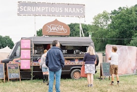 A black food truck adorned with colorful patterns is parked in a grassy area with a large sign on top that reads 'SCRUMPTIOUS NAANS'. Several chalkboards display menu options, including breakfast naan and juices. Three people are standing near the truck, likely customers waiting to order. Trees and tents are visible in the background.