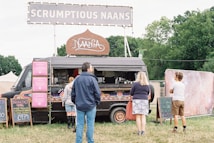 A black food truck adorned with colorful patterns is parked in a grassy area with a large sign on top that reads 'SCRUMPTIOUS NAANS'. Several chalkboards display menu options, including breakfast naan and juices. Three people are standing near the truck, likely customers waiting to order. Trees and tents are visible in the background.