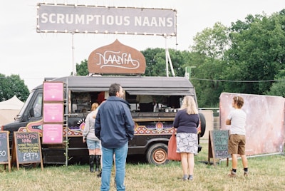 A black food truck adorned with colorful patterns is parked in a grassy area with a large sign on top that reads 'SCRUMPTIOUS NAANS'. Several chalkboards display menu options, including breakfast naan and juices. Three people are standing near the truck, likely customers waiting to order. Trees and tents are visible in the background.