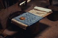 A wooden surface with intricately designed fabric laid out. The fabric features detailed geometric patterns, including a prominent blue piece with white circular motifs. A small brown item, resembling a roll or loaf, rests on the fabric, next to a rolled-up piece of material. The setting appears rustic, with a woven basket partially visible.
