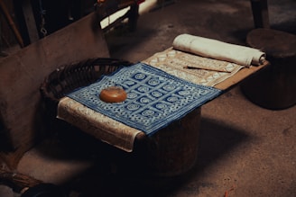 A wooden surface with intricately designed fabric laid out. The fabric features detailed geometric patterns, including a prominent blue piece with white circular motifs. A small brown item, resembling a roll or loaf, rests on the fabric, next to a rolled-up piece of material. The setting appears rustic, with a woven basket partially visible.
