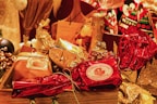 A festive arrangement featuring wrapped gifts in various metallic and festive colors, including red and gold. The centerpiece is a wrapped item labeled 'Authentic Panettone.' Surrounding it are other wrapped gifts and a bottle of champagne, creating a celebratory and luxurious atmosphere.