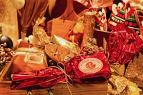 A festive arrangement featuring wrapped gifts in various metallic and festive colors, including red and gold. The centerpiece is a wrapped item labeled 'Authentic Panettone.' Surrounding it are other wrapped gifts and a bottle of champagne, creating a celebratory and luxurious atmosphere.