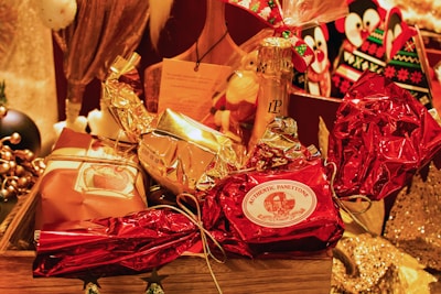 A festive arrangement featuring wrapped gifts in various metallic and festive colors, including red and gold. The centerpiece is a wrapped item labeled 'Authentic Panettone.' Surrounding it are other wrapped gifts and a bottle of champagne, creating a celebratory and luxurious atmosphere.