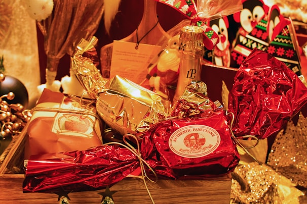 A festive arrangement featuring wrapped gifts in various metallic and festive colors, including red and gold. The centerpiece is a wrapped item labeled 'Authentic Panettone.' Surrounding it are other wrapped gifts and a bottle of champagne, creating a celebratory and luxurious atmosphere.