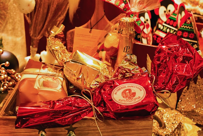 A festive arrangement featuring wrapped gifts in various metallic and festive colors, including red and gold. The centerpiece is a wrapped item labeled 'Authentic Panettone.' Surrounding it are other wrapped gifts and a bottle of champagne, creating a celebratory and luxurious atmosphere.