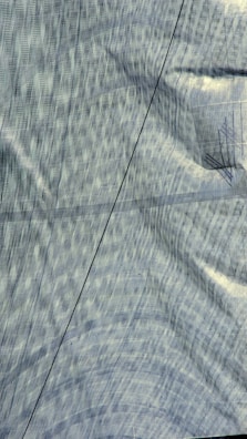 A textured fabric surface with a prominent crease running diagonally across it. The fabric displays a subtle, interwoven pattern with a mix of straight and curved lines. Lighting effects highlight the fabric's structure, creating a sense of depth and movement.