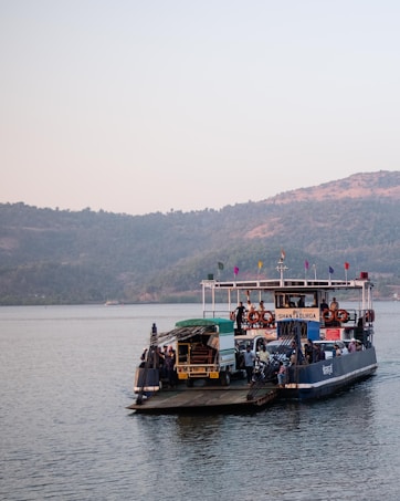 A ferry carrying vehicles and people is moving across a body of water. The ferry is named Shantadurga and is equipped with safety lifebuoys. The surrounding landscape includes hills covered in greenery and a calm, clear sky.