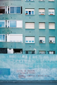 A teal-colored apartment building with multiple floors and closed windows. The lower part is a wall with red graffiti that includes political messages mentioning countries like Colombia, Morocco, and Palestine. The scene depicts an urban area.