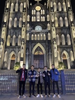A group of five people stand in front of a large, intricately designed Gothic-style cathedral illuminated at night. The facade features pointed arches, numerous windows, and decorative stonework. Above, the word 'Emmanuel 2022' is displayed.