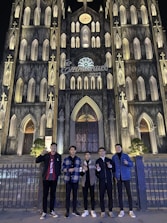 A group of five people stand in front of a large, intricately designed Gothic-style cathedral illuminated at night. The facade features pointed arches, numerous windows, and decorative stonework. Above, the word 'Emmanuel 2022' is displayed.