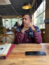 A person sits in a cozy, modern cafe with wooden tables and large windows allowing natural light. The individual appears contemplative, with hands clasped near their face. There are hanging lamps, and a menu is visible on the table.