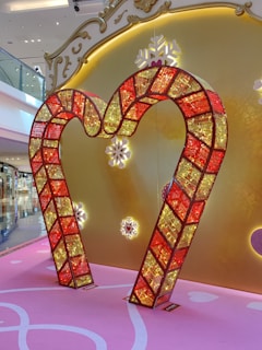 A decorative frame in the shape of a heart made from red and yellow illuminated panels with a mosaic-like design is displayed against a backdrop featuring a golden ornate pattern with snowflake motifs. The floor is pink with white accents, contributing to a festive and elegant ambience.