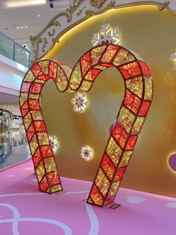 A decorative frame in the shape of a heart made from red and yellow illuminated panels with a mosaic-like design is displayed against a backdrop featuring a golden ornate pattern with snowflake motifs. The floor is pink with white accents, contributing to a festive and elegant ambience.