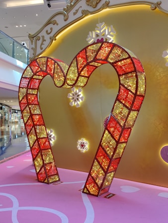 A decorative frame in the shape of a heart made from red and yellow illuminated panels with a mosaic-like design is displayed against a backdrop featuring a golden ornate pattern with snowflake motifs. The floor is pink with white accents, contributing to a festive and elegant ambience.