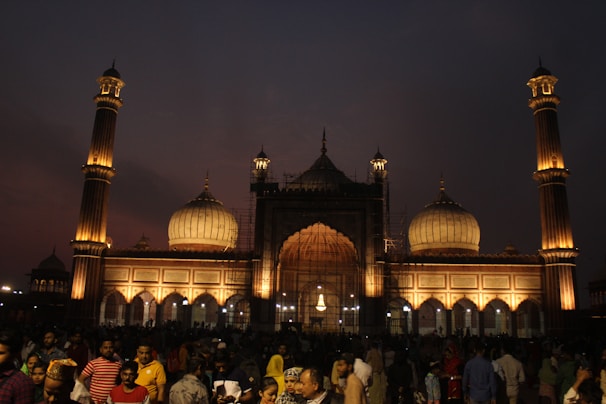 A large crowd gathers in front of a grand mosque illuminated by warm lights against a dusky sky. The structure features multiple domes and tall minarets, showcasing intricate architectural details. People of various ages are seen mingling and socializing in the foreground, indicating a lively atmosphere.
