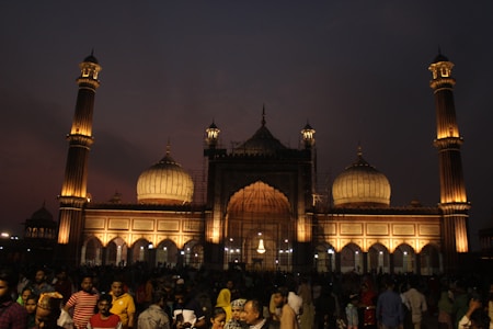 A large crowd gathers in front of a grand mosque illuminated by warm lights against a dusky sky. The structure features multiple domes and tall minarets, showcasing intricate architectural details. People of various ages are seen mingling and socializing in the foreground, indicating a lively atmosphere.