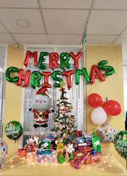 A festive Christmas display in an office setting featuring a decorated Christmas tree with ornaments and a star on top. Below the tree, various brightly wrapped gifts are arranged on a table. The backdrop includes metallic streamers and a large Santa balloon decoration. Above the tree, red and green balloons spell out 'MERRY CHRISTMAS'. Additional balloons with festive designs are placed around the setup, contributing to the cheerful atmosphere.