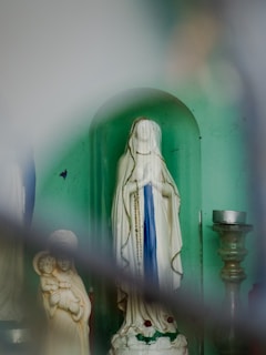A religious setting featuring a statue of the Virgin Mary adorned in a white robe with blue accents, enclosed in a small alcove. Adjacent to the main statue is a smaller statue of another figure holding a child. A candle holder is visible to the right. The setting gives a sense of a prayer or meditation space.