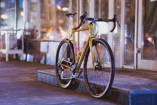 A sleek, modern bicycle with a vibrant yellow frame is parked on a sidewalk next to a building with large glass windows. The setting appears to be an urban environment with softly blurred lights in the background, suggesting a possibly busy street or city setting during the evening.